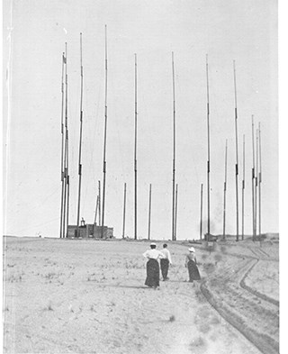 A black and white photo of a man and two women standing in an open area facing a building next to a tall circular array of thin antennas. 